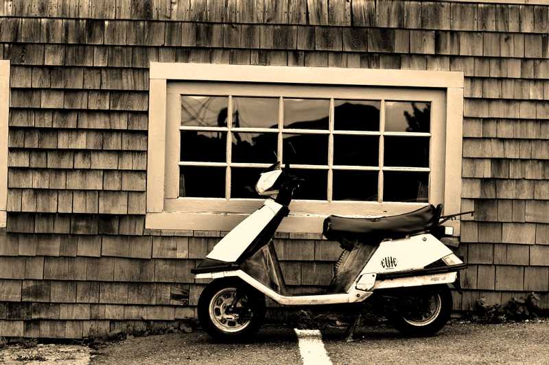 A black and white shot of a moped parked in front of a big restaurant window.