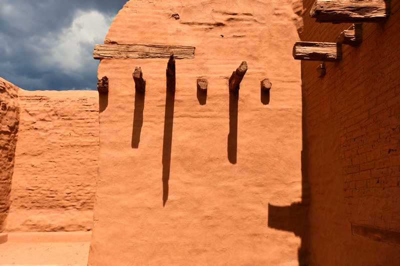 A wall of a pueblo with long shadows cast from the supporting wooden beams